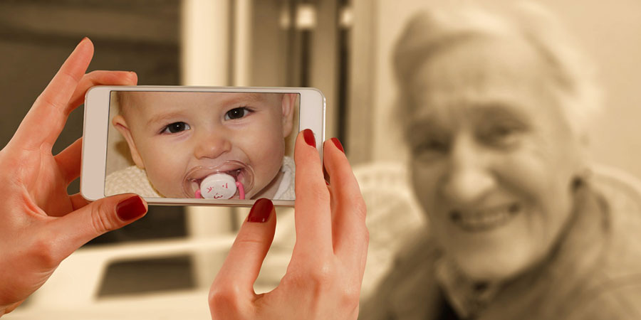 kädet pitävät puhelinta, jossa vauvan kuva, taustalla hymyilevä ikäihminen.