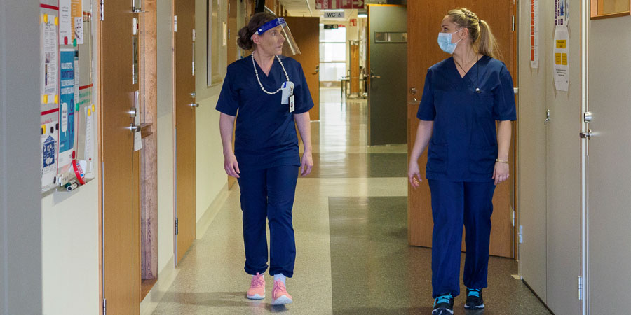Kaksi henkilöä, joilla sairaalan henkilökunnan vaatetus, kävelee sisäkäytävää, jonka seinillä useita ovia ja ilmoitustauluja.