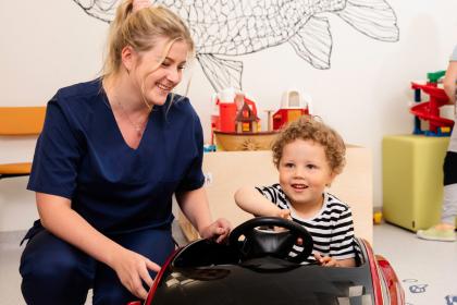 A child sits on a toy car. A nurse is beside the child.