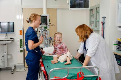 Hoitaja ja lääkäri tutkivat lasta, joka istuu sairaalasängyllä.