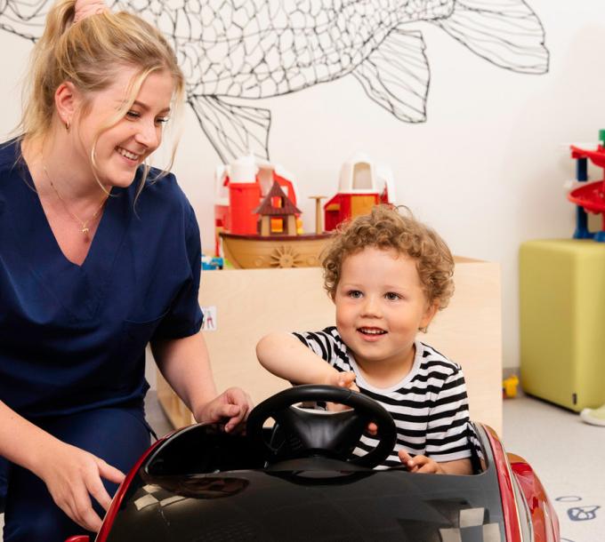 A child sits on a toy car. A nurse is beside the child.