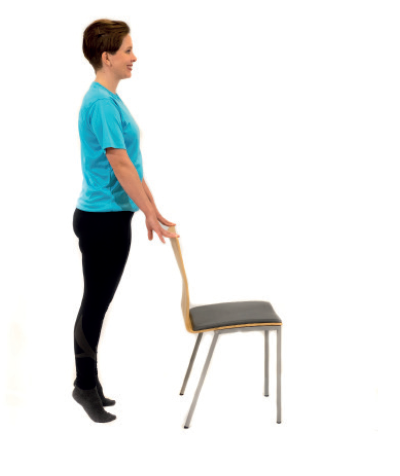 nainen seisoo varpaillaan pitäen tuolin selkänojasta kiinni.